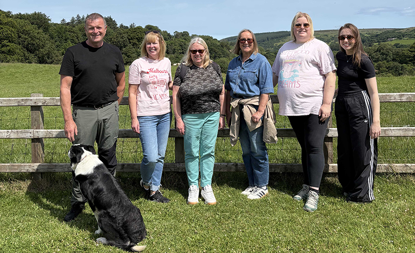 Group of six adults and a black-and-white dog standing by a wooden fence in a sunny green countryside landscape.