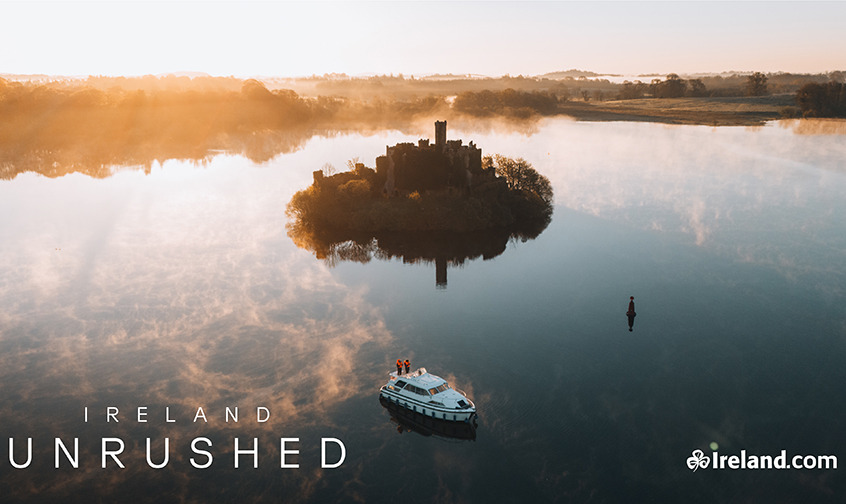 Aerial view of a small castle island reflected in calm water at sunrise, with a boat passing nearby