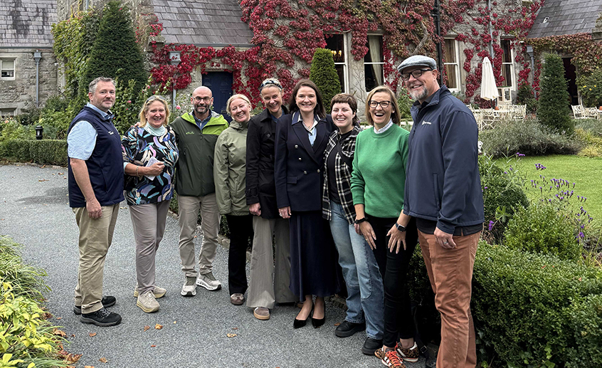 Group of eight people standing outside a stone building covered in red ivy, posing together on a pathway in a landscaped garden.