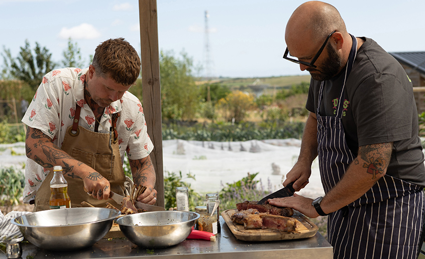 Two chefs preparing and carving food outdoors in a garden setting.