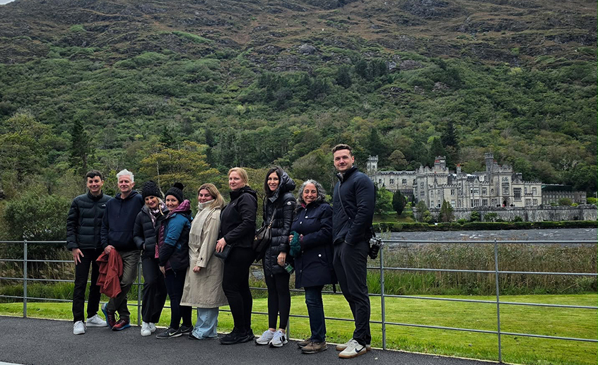 Group of travel agents posing outdoors with a historic castle and mountainous landscape in the background.