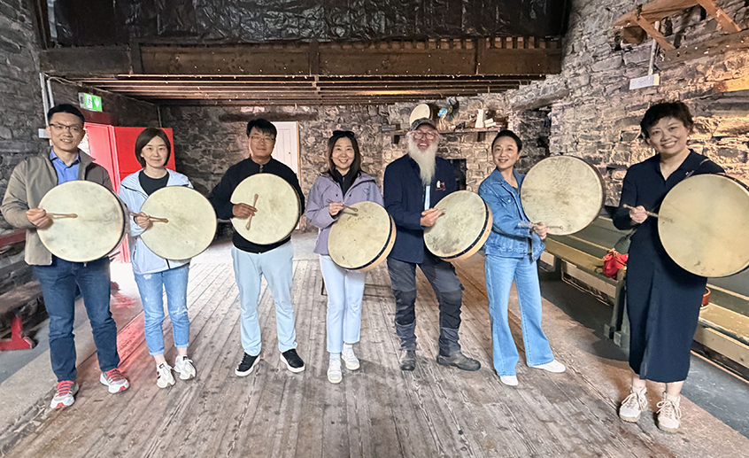 Group of seven people holding traditional frame drums inside a rustic stone building with wooden floors and exposed beams.