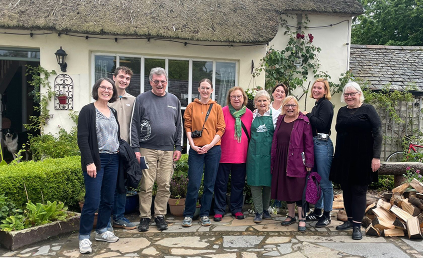 Group of people standing outside a traditional thatched cottage, posing together in a garden setting with stone paving and flowers.