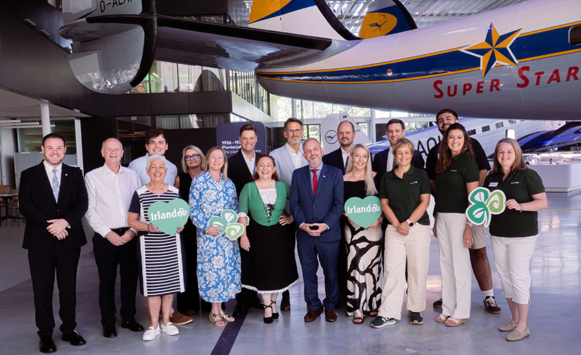 Large group of adults posing inside an aviation museum beneath a vintage “Super Star” aircraft, some holding Ireland-branded signs.