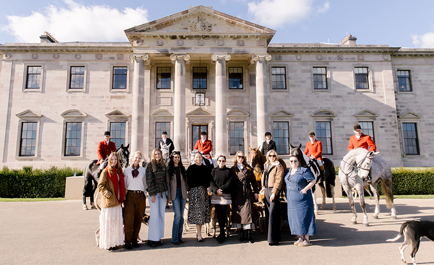 Group of visitors and horse riders posing in front of a grand historic country house on a sunny day.