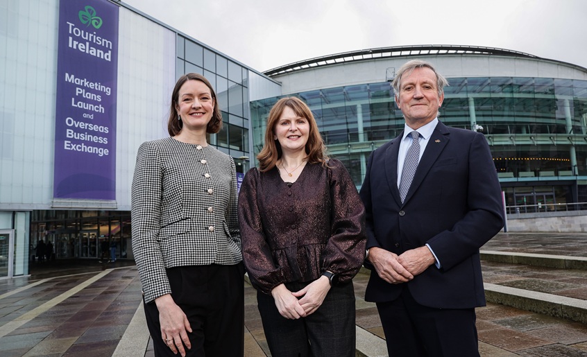 Three people standing outside a dome ceilinged building with a purple Tourism Ireland banner saying 'Tourism Ireland Marketing Plans Launch and Overseas Business Exchange'