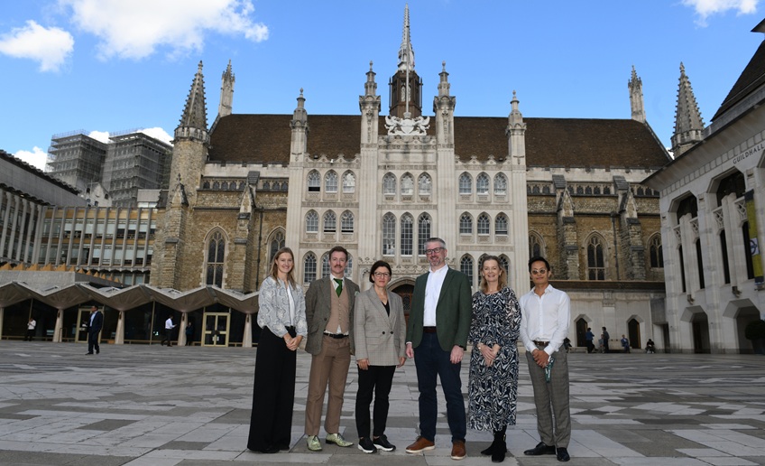 Six people stand in a city square in front of a large historic Gothic-style building under a bright blue sky.