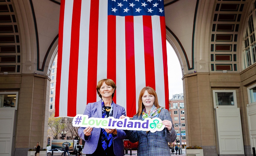 Two women standing in front of an archway with the US flag in the background, holding a 'Love Ireland' sign.