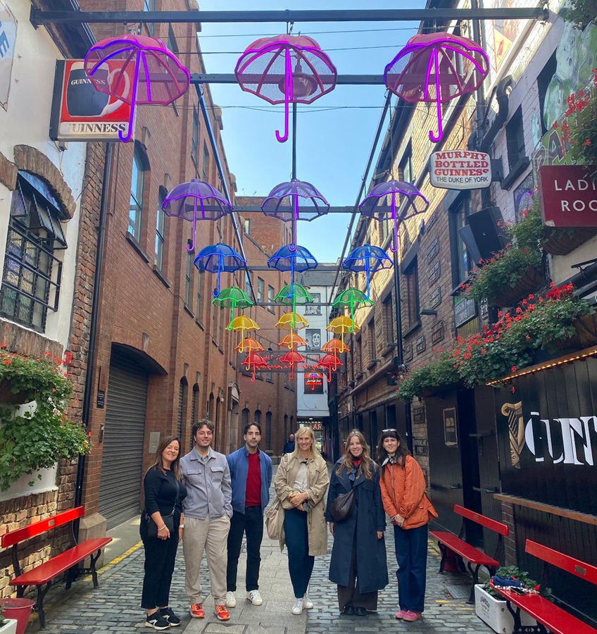   Six people standing in a cobbled alley beneath colourful illuminated umbrella decorations, with brick buildings, pub signs and flower boxes lining the street.