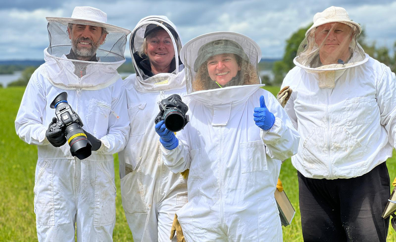 Four people in white beekeeping suits standing in a grassy field, holding cameras.