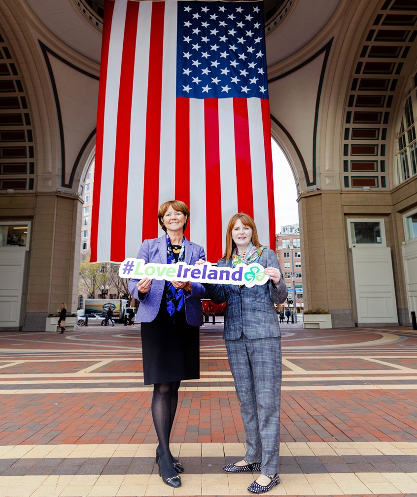Two women standing in front of an archway with the US flag in the background, holding a 'Love Ireland' sign. 