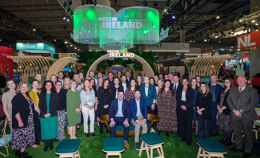 Group photo of a large team standing in front of a green-themed ‘Meet in Ireland’ booth at a trade show, featuring wooden arches, greenery, and illuminated signage
