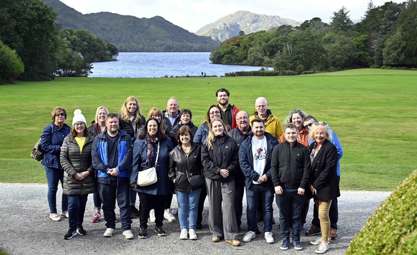 A group of people standing together outdoors in hotel grounds with a lake and mountains behind them.
