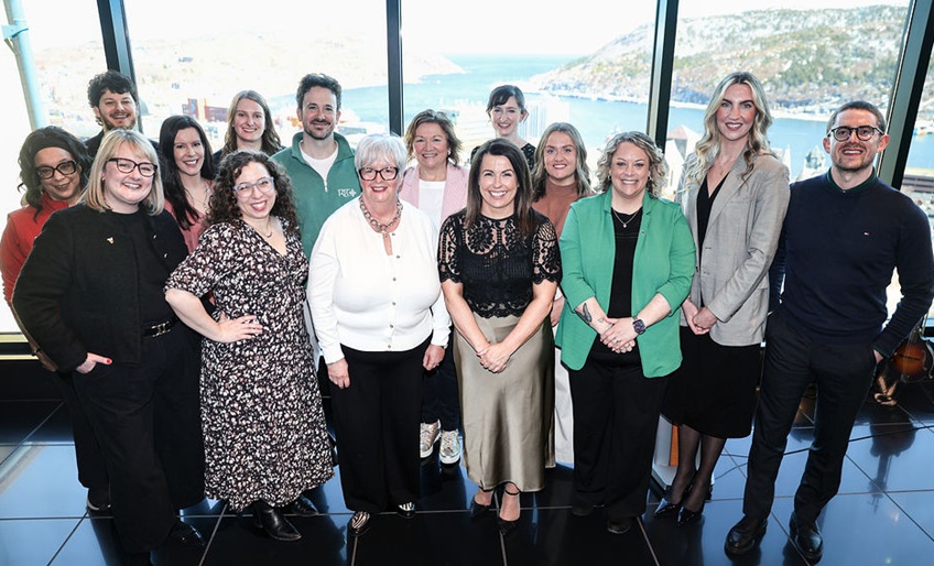 Indoor picture of group of people standing in front of a window with scenic mountain and water outside.