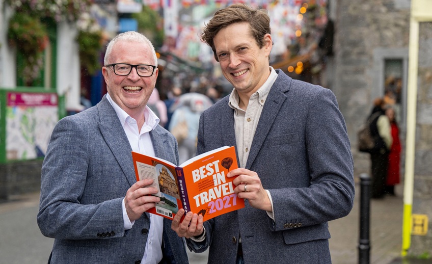 Two people standing on a street, holding and looking at an orange travel guidebook titled “Best in Travel 2024,” with a blurred town streetscape in the background.