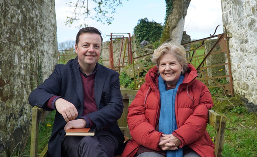 Two people seated outdoors in a rural setting, with stone walls and countryside in the background.